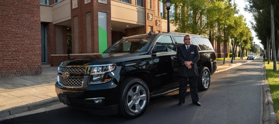 A uniformed chauffeur standing beside a black vehicle on a tree-lined street.